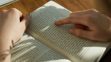 Hands holding a book open with a finger pointing to the text.