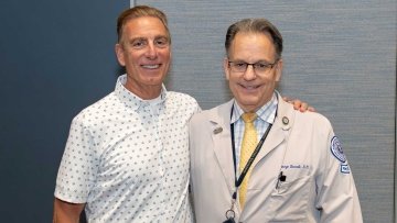 Mr. Giglio and Dr. Borelli in a white coat and stethoscope take a photo together at the patient panel.