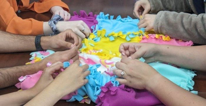 Students work together to make no-sew blankets. Five pairs of hands make the no-sew blankets in pink, yellow, turquoise, and purple material.